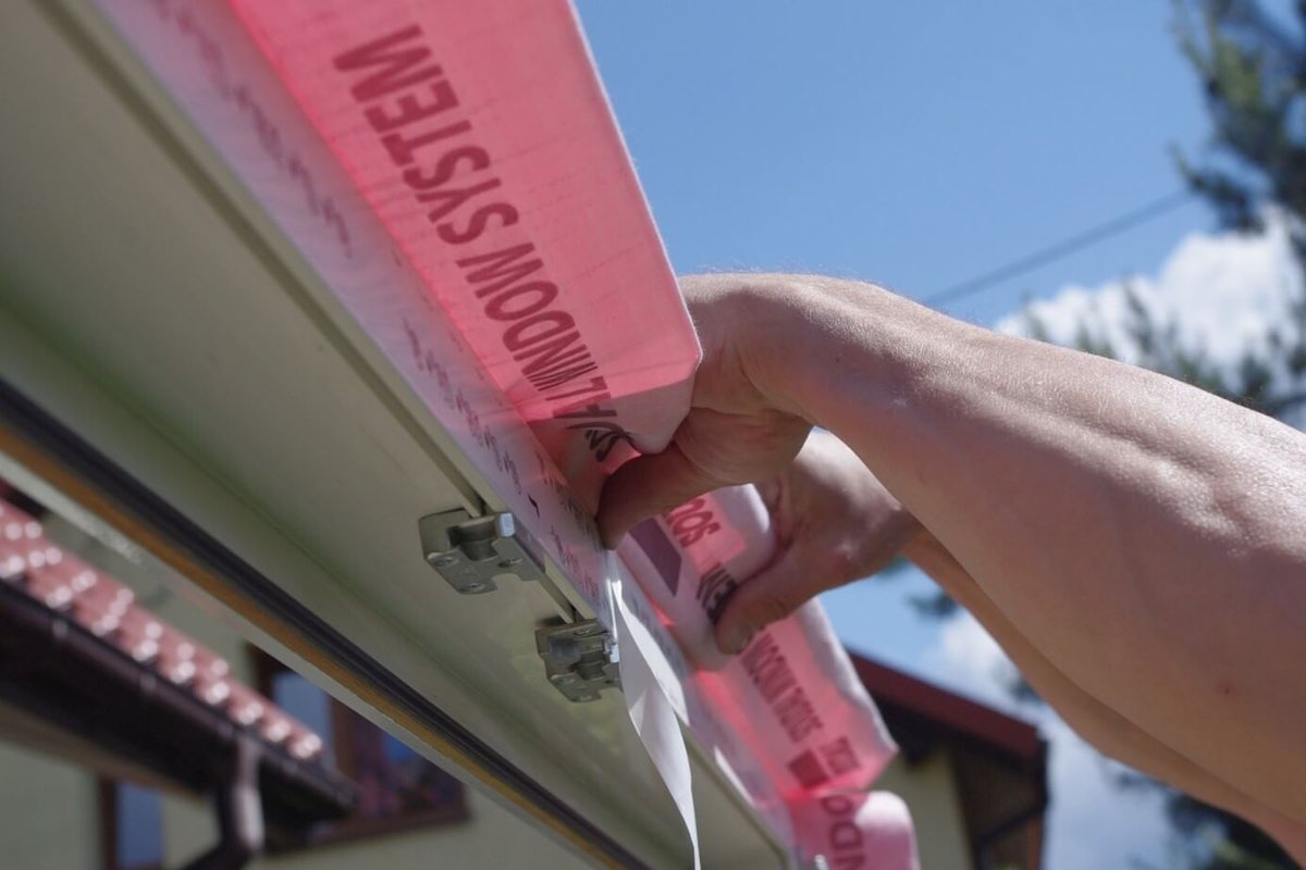 Dłoń przykleja różową taśmę paroszczelną do krawędzi ramy okna PVC, na ramie widać stalowe kotwy